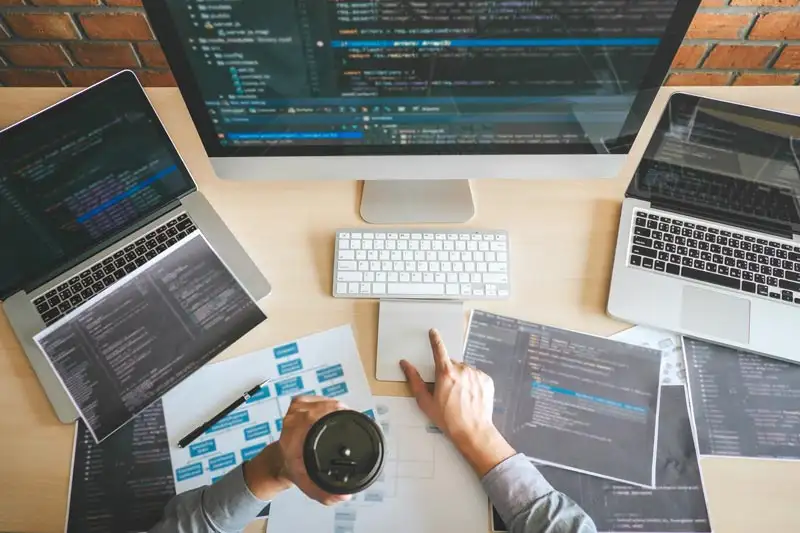 Aerial view of a programmer working on multiple screens with coding documents and a coffee cup on the desk.